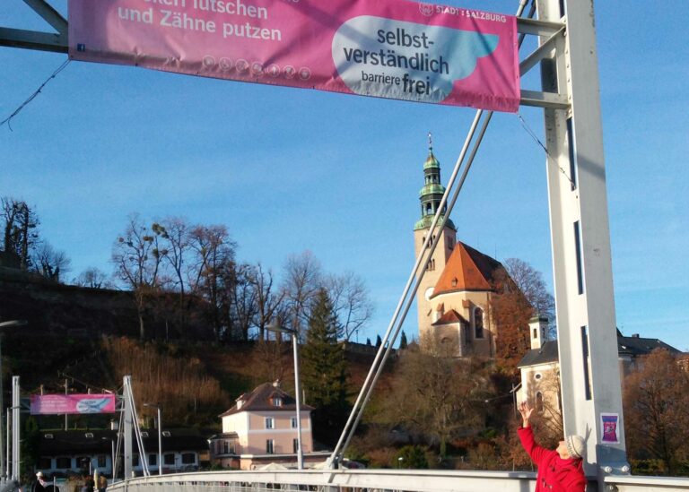 On a pedestrian bridge over a river, a person in a red coat points to a banner with the inscription "self-evidently accessible, City of Salzburg." In the background, there is an old town area with a church and historic building