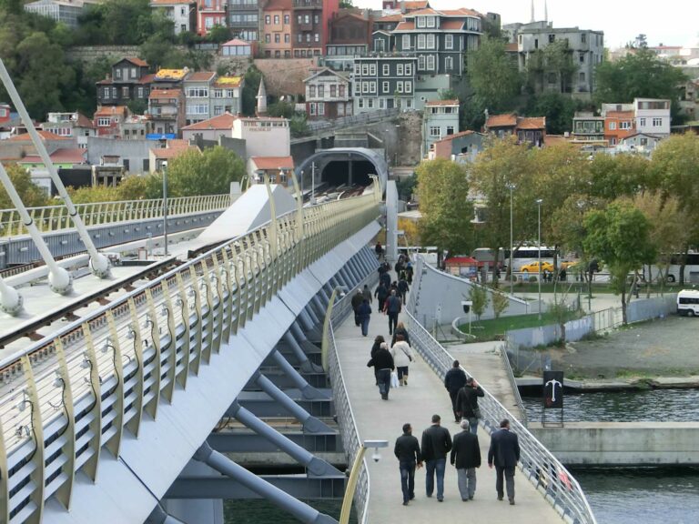 A modern bridge, consisting of a subway bridge with an attached curved pedestrian bridge filled with many people, leads over a canal into a dense city on a hillside